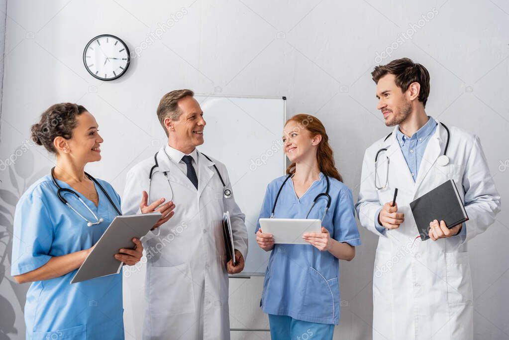 Happy multicultural doctors and nurses looking at each other while standing together with flipchart and wall clock on background