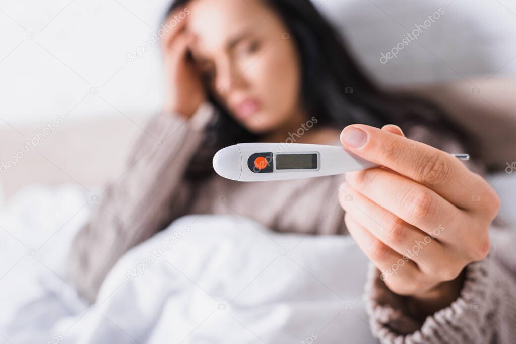 sick young brunette woman showing thermometer in bed, blurred background