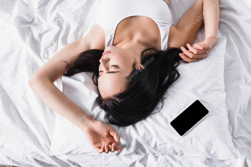 top view of young brunette woman sleeping in bed near smartphone