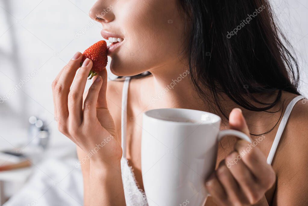 cropped view of young brunette woman with mug eating strawberry for breakfast