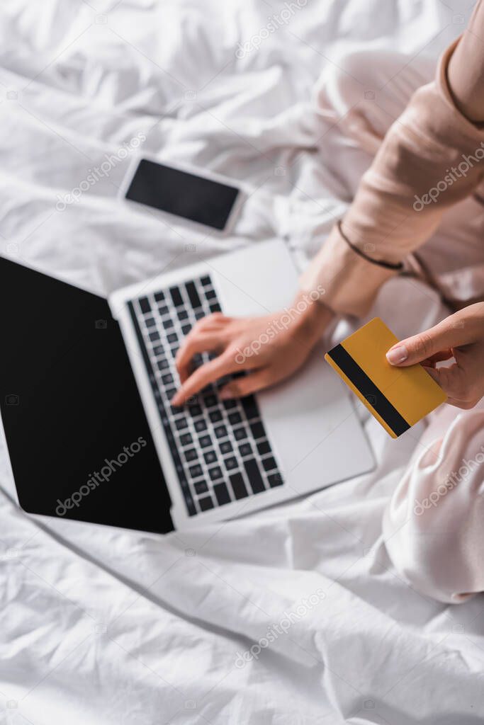 cropped view of woman sitting in bed with smartphone, credit card and laptop at morning