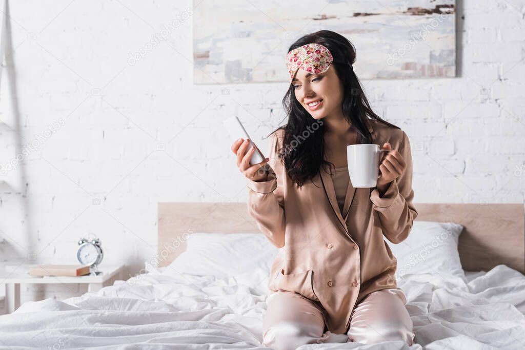 smiling young brunette woman sitting in bed in sleep mask with mug of cocoa and smartphone