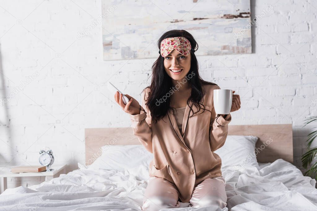 smiling young brunette woman sitting in bed in sleep mask with mug of cocoa and smartphone