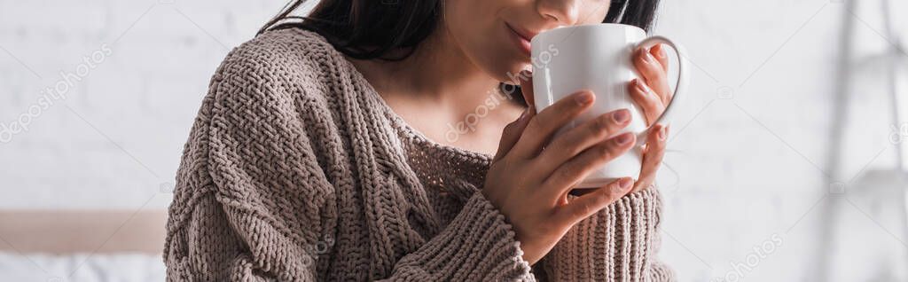 cropped view of young brunette woman in sweater with mug of hot cocoa at morning, banner