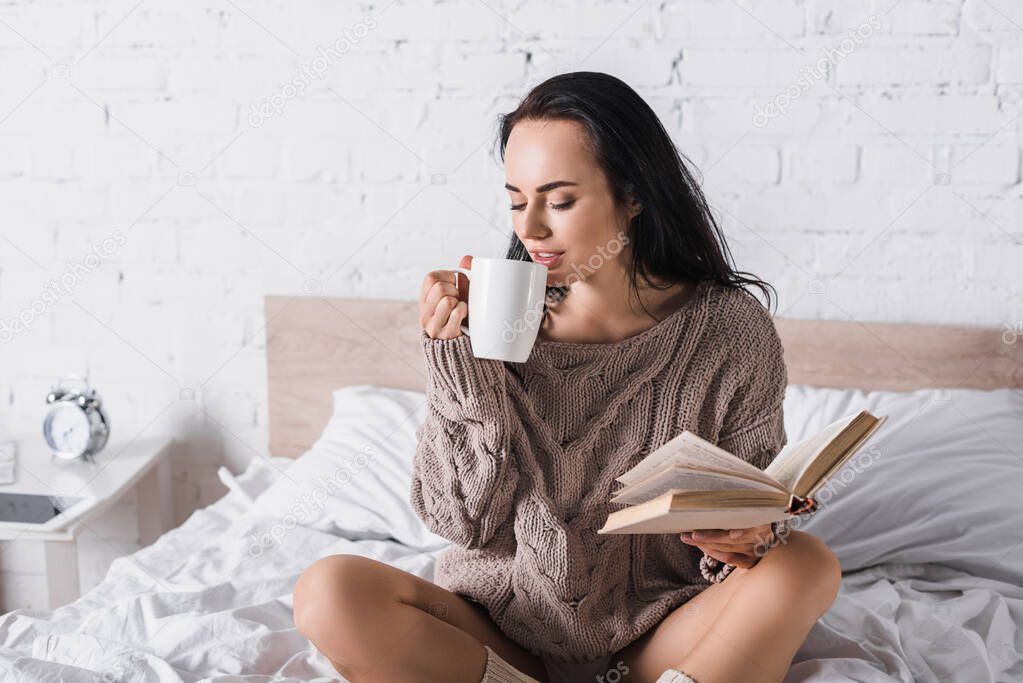 young brunette woman in sweater sitting in bed with mug of hot cocoa and book at morning
