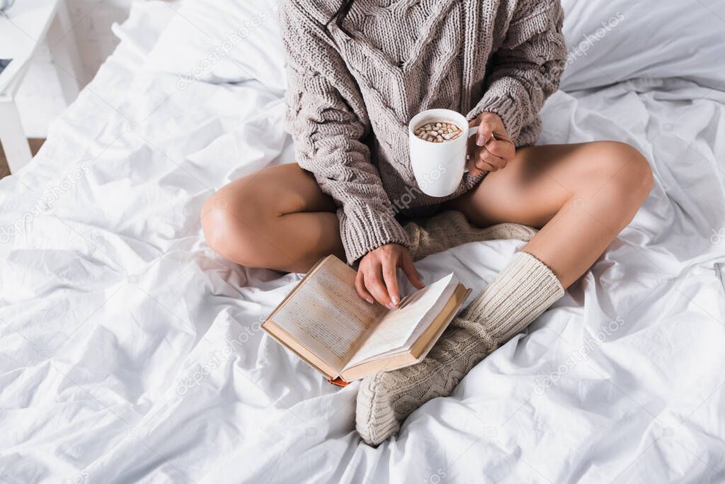 cropped view of woman in sweater and knitted socks sitting in bed with mug of hot cocoa and book at morning