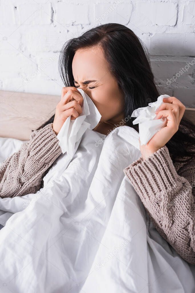 ill young brunette woman in sweater sneezing in bed with tissue