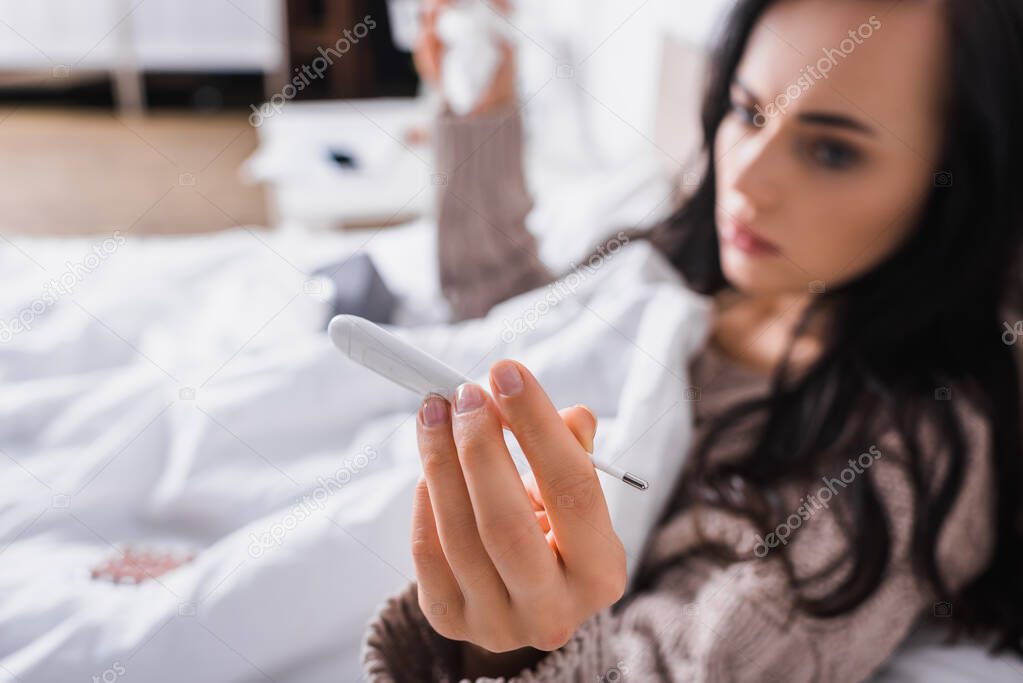 blurred ill young brunette woman in sweater lying in bed with thermometer and tissue