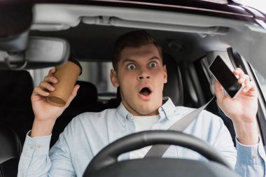 scared man holding coffee to go and smartphone at steering wheel in car, blurred foreground clipart