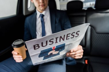 Cropped view of newspaper in hand of businessman with coffee to go in car on blurred background clipart
