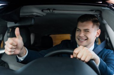 Smiling businessman winking and showing like gesture while driving car on blurred foreground clipart
