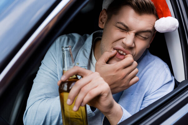 пьяный мужчина в шляпе Санты свистит, сидя в машине с бутылкой виски, размытый передний план
