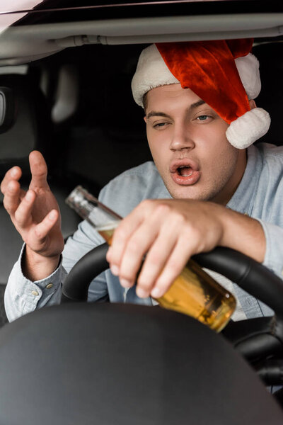 пьяный мужчина в шляпе Санты держит бутылку алкоголя и жестикулирует во время вождения автомобиля на размытом переднем плане