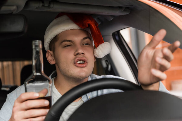 недовольный пьяный мужчина в шляпе Санты жестом сидя за рулем с бутылкой виски, размытый передний план