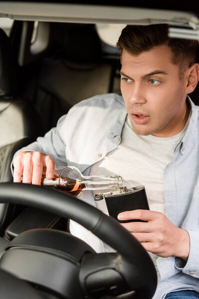 пьяный человек разливает виски из бутылки в фляжку, сидя в машине, размытый передний план