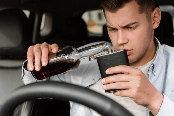 пьяный молодой человек разливает виски из бутылки в фляжку в машине на размытом переднем плане