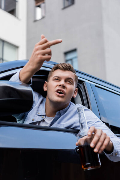 пьяный, агрессивный человек с бутылкой виски показывая средний палец, глядя в окно машины, размытый передний план