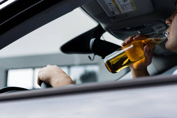 частичный вид человека, пьющего виски во время вождения автомобиля на размытом переднем плане
