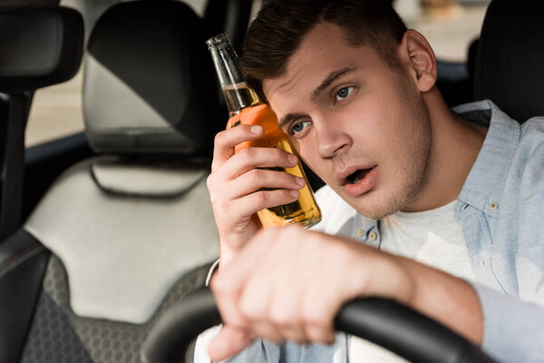 пьяный мужчина держит бутылку алкоголя возле головы во время вождения автомобиля на размытом переднем плане