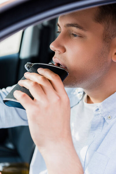 Молодой человек пьет алкоголь из фляжки во время вождения автомобиля, размытый передний план