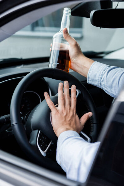частичный вид человека, пищащего во время вождения автомобиля и держащего бутылку виски, размытый передний план