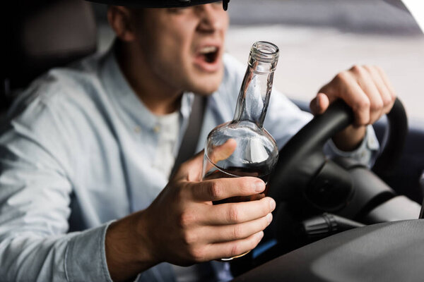 обрезанный вид агрессивного пьяного человека с бутылкой виски, кричащего во время вождения автомобиля