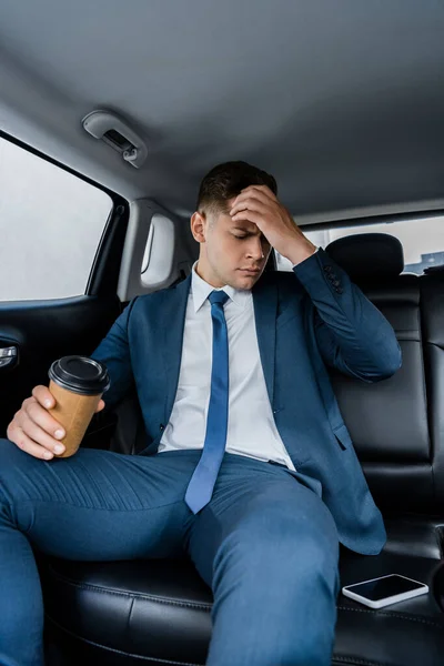 疲惫的商人拿着外卖咖啡广告 看着车内装有空白屏幕的智能手机 — 图库照片