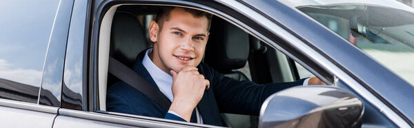 Smiling businessman with hand near chin looking at camera in auto, banner