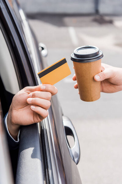 Cropped view of man holding credit card in auto near seller with takeaway coffee