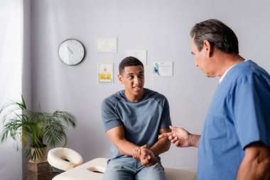 chiropractor pointing with finger at injured arm of african american patient clipart