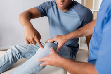cropped view of african american patient pointing with finger at knee near doctor  clipart