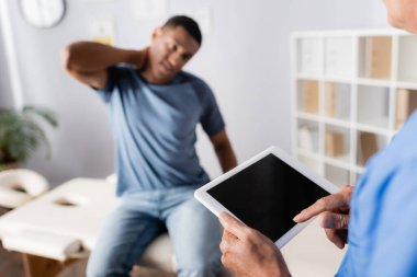 Olgun doktor, arka planda bulanık boyun ağrısından muzdarip Afrikalı Amerikalı bir hastanın yanında dijital tablet kullanıyor. 