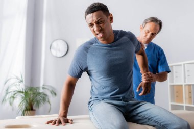 injured african american man suffering from pain near masseur on blurred background clipart