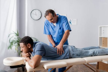 Orta yaşlı kayropraktik uzmanı, Afrikalı Amerikalı bir hastanın sırtında, klinikteki masaj masasında çalışıyor.