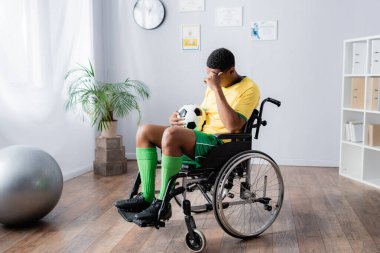 Tekerlekli sandalyedeki özürlü Afro-Amerikan sporcusu elinde futbol topu tutuyor. 