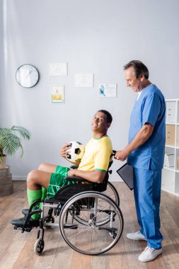 Tekerlekli sandalyedeki neşeli Afro-Amerikan sporcusu elinde futbol topu ve doktorun yanında gülümsüyor. 