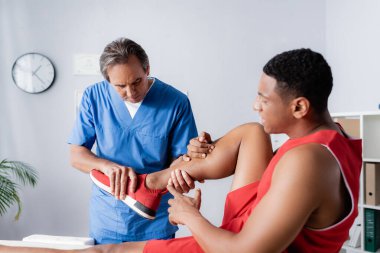 Spor ayakkabılı Afrikalı Amerikalı bir adam klinikteki kayropraktik uzmanının yanındaki yaralı bacağı işaret ediyor.