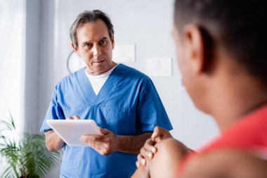 doctor with digital tablet looking at african american man on blurred foreground clipart