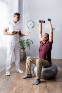 Afrika kökenli Amerikalı fizyoterapistin yanında elinde dijital tabletle spor salonunu işaret eden halterlerle spor yapan orta yaşlı bir adam.
