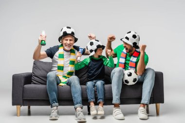 Dede ve babasıyla heyecanlı çocuk gri şapkalarla futbol maçı izlerken zafer jesti yapıyor.