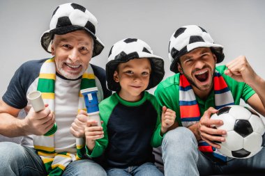 Heyecanlı adam çığlık atıyor ve galibiyet işareti yapıyor. Futbol fanatik şapkalı baba ve oğlunun yanında. Gri renkte izole edilmiş.