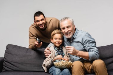 Baba ve oğlunun yanında uzaktan kumandayı tutan mutlu bir adam televizyon izliyor ve grinin üzerinde izole edilmiş patlamış mısır yiyor.
