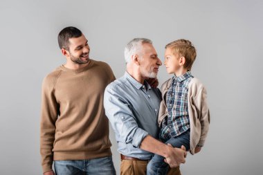 happy mature man holding grandson near smiling son isolated on grey clipart