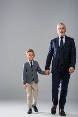 mature businessman holding hand of grandson while walking on grey clipart