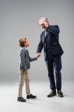 happy businessman touching eyeglasses while holding hands with grandson on grey clipart