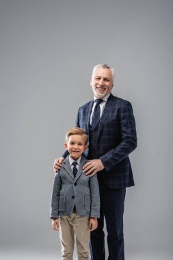 happy businessman in formal wear hugging shoulders of grandson while looking at camera isolated on grey clipart