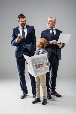 smiling boy with newspaper near dad and grandfather holding gadgets on grey clipart