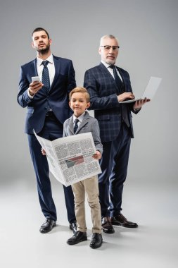 Babam ve büyükbabamın yanında kameraya bakan, elinde gri aletlerle gülümseyen bir çocuk.