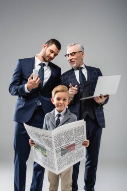 mature businessman talking to son and pointing with finger at grandson holding newspaper on grey clipart