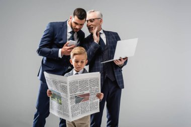 mature businessman talking to son near boy in formal wear reading newspaper isolated on grey clipart